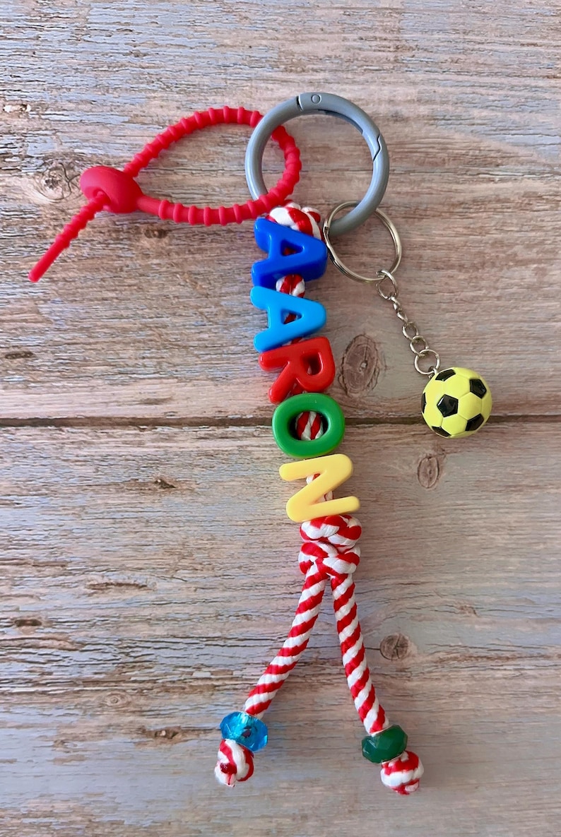 Op de afbeelding: Een gepersonaliseerde sleutelhanger met de naam "AARON" in kleurrijke letterkralen. De sleutelhanger bevat een voetbalbedel en een rood-wit gestreept touwontwerp. Een zilverkleurige sleutelring en een rode siliconen lus maken het ontwerp compleet.