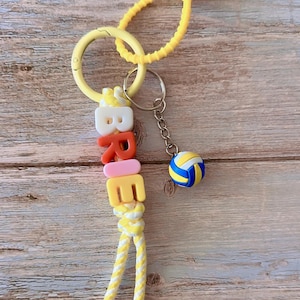 Op de afbeelding: Een gele sleutelhanger met de naam "BREE" in kleurrijke letterkralen. Een kleine volleybalbedel hangt aan de sleutelhanger. De sleutelhanger heeft een gele ring en een geel koord.