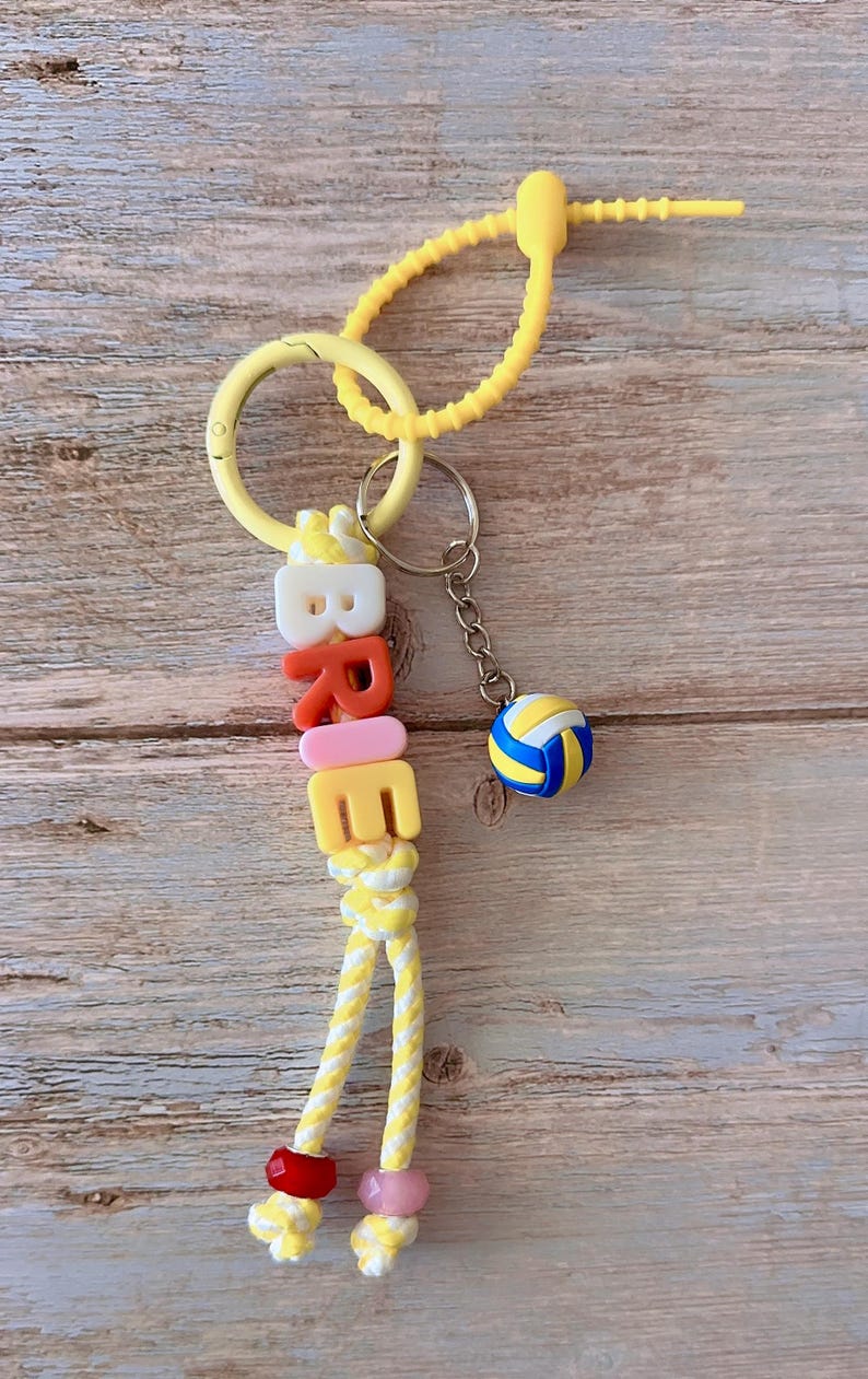 Op de afbeelding: Een gele sleutelhanger met de naam "BREE" in kleurrijke letterkralen. Een kleine volleybalbedel hangt aan de sleutelhanger. De sleutelhanger heeft een gele ring en een geel koord.