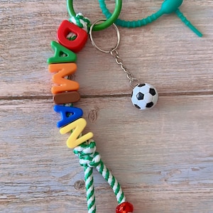 Op de afbeelding: Een kleurrijke sleutelhanger met de naam "DAMIAN" gespeld in individuele letterkralen. De sleutelhanger bevat een kleine voetbalbedel en een groen en wit touwdetail. Hij is bevestigd aan een groene sluiting en een turquoise rubberen lus.