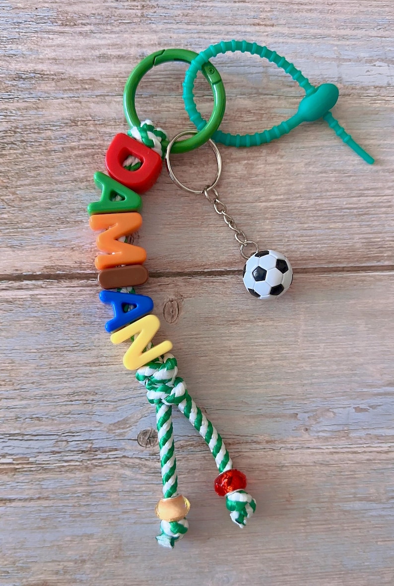 Op de afbeelding: Een kleurrijke sleutelhanger met de naam "DAMIAN" gespeld in individuele letterkralen. De sleutelhanger bevat een kleine voetbalbedel en een groen en wit touwdetail. Hij is bevestigd aan een groene sluiting en een turquoise rubberen lus.