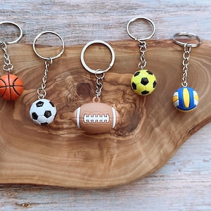 Op de afbeelding: Sleutelhangers met sportthema's, met miniatuur honkbal, basketbal, voetbal, American football en volleybal ontwerpen. Elke sleutelhanger heeft een zilverkleurige ring en ketting, gepresenteerd op een houten oppervlak.