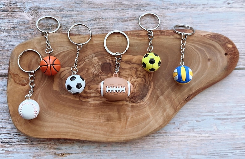 Op de afbeelding: Sleutelhangers met sportthema's, met miniatuur honkbal, basketbal, voetbal, American football en volleybal ontwerpen. Elke sleutelhanger heeft een zilverkleurige ring en ketting, gepresenteerd op een houten oppervlak.