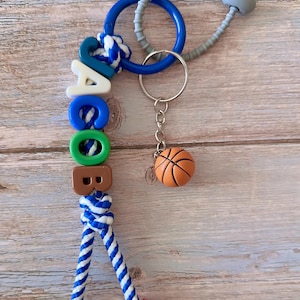 Op de afbeelding: Een gepersonaliseerde sleutelhanger met de naam "JACOB" gespeld met kleurrijke letterkralen. De sleutelhanger bevat een kleine basketbalbedel en een blauw-wit touwontwerp. De achtergrond is een licht houten oppervlak.