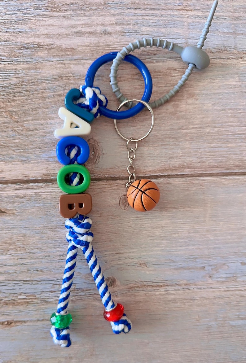 Op de afbeelding: Een gepersonaliseerde sleutelhanger met de naam "JACOB" gespeld met kleurrijke letterkralen. De sleutelhanger bevat een kleine basketbalbedel en een blauw-wit touwontwerp. De achtergrond is een licht houten oppervlak.