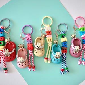 May include: A collection of colorful keychains featuring miniature shoe charms. Each keychain has a different color shoe, a small bell, and a name spelled out with letter beads. The keychains are arranged on a turquoise and white background.