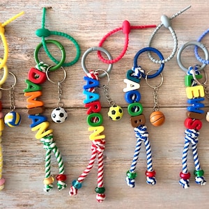 Op de afbeelding: Gepersonaliseerde sleutelhangers met namen geschreven in kleurrijke alfabetkralen. Elke sleutelhanger heeft een sportthema-bedel, zoals een voetbal, basketbal of rugbybal. De sleutelhangers liggen op een houten oppervlak.