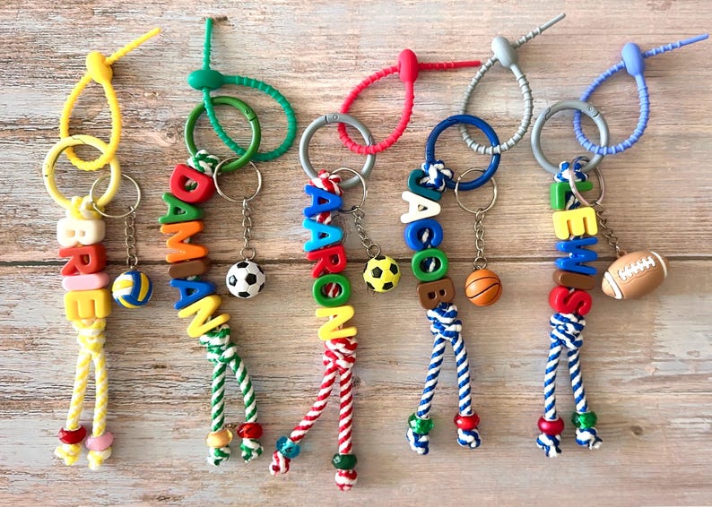 Op de afbeelding: Gepersonaliseerde sleutelhangers met namen geschreven in kleurrijke alfabetkralen. Elke sleutelhanger heeft een sportthema-bedel, zoals een voetbal, basketbal of rugbybal. De sleutelhangers liggen op een houten oppervlak.
