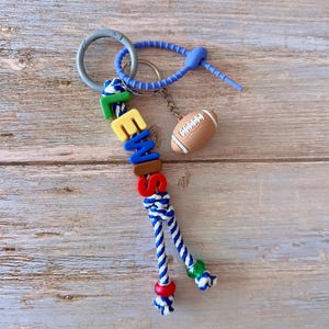 Op de afbeelding: Een gepersonaliseerde sleutelhanger met de naam "EMME" in kleurrijke letterkralen. De sleutelhanger heeft een blauw-wit touwontwerp, een voetbalbedel en een zilveren sluiting.