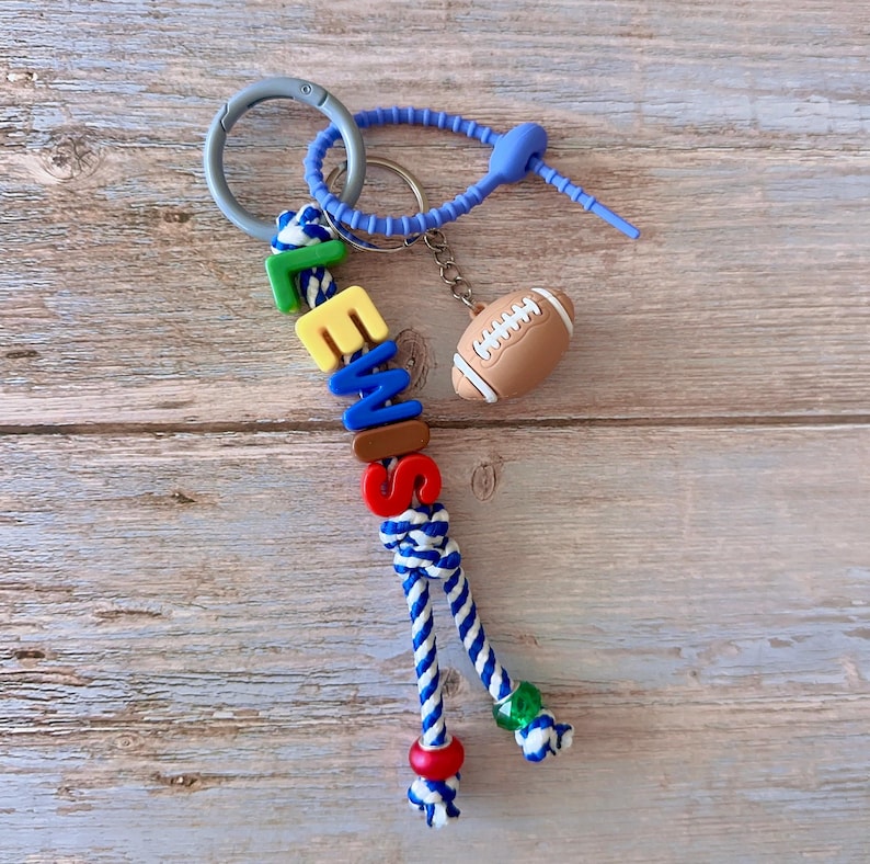 Op de afbeelding: Een gepersonaliseerde sleutelhanger met de naam "EMME" in kleurrijke letterkralen. De sleutelhanger heeft een blauw-wit touwontwerp, een voetbalbedel en een zilveren sluiting.