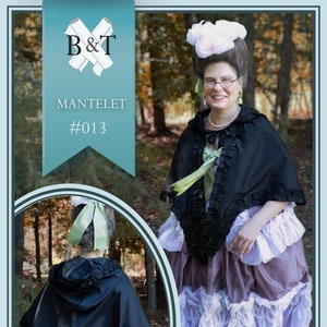 May include: A black hooded cape with a ruffled trim, worn over a brown and white ruffled skirt. The cape is styled with a green ribbon tied at the back of the neck. The image includes the text "MANTELET #013".