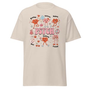 May include: A beige t-shirt featuring a central graphic with the word PSYCH in large pink letters. The design includes various cartoon heart characters, a disco ball, a teddy bear, cherries, and text labels including Meetings, Referrals, Reports, Data, Positivity, and Feelings.