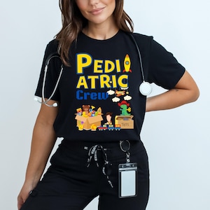Może przedstawiać: Czarny T-shirt z napisem "Pediatric Crew" w żółtych i niebieskich literach. Koszulka przedstawia kreskówkową ilustrację pudełka wypełnionego zabawkami, w tym pluszowym misiem, rakietą, dinozaurem i ciężarówką.