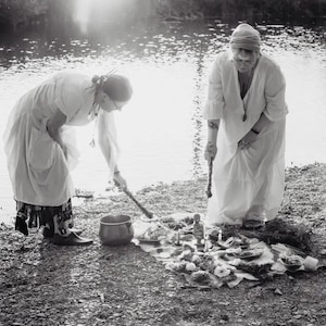 Könnte beinhalten: Zwei Personen in weißen Roben stehen an einem Fluss und führen ein Ritual mit einem Kessel und verschiedenen Gegenständen auf einer Decke durch. Die Szene ist in Schwarzweiß.
