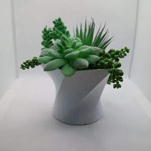 May include: A white, geometric-shaped planter filled with a variety of artificial succulents. The arrangement includes a large, central succulent with green leaves, smaller round succulents, and spiky green plants. The planter is approximately 15 cm tall.