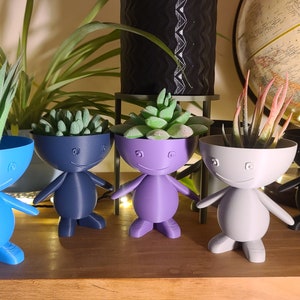 May include: Five small, colourful, 3D printed planters shaped like people with smiling faces. Each planter has a different colour: blue, dark blue, purple, grey, and black. The planters are filled with small plants.
