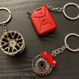 May include: Three silver keychains with different car-themed charms. One keychain has a silver rimmed wheel, another has a red gas can with the text "20L" on it, and the third has a red brake rotor with a red car on top.
