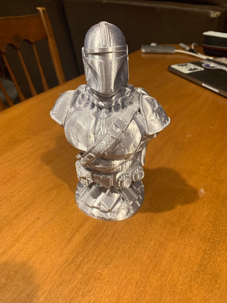 Puede incluir: Busto plateado de un personaje mandaloriano, con casco, armadura de hombro y peto con cintur&oacute;n y bolsas. El busto est&aacute; impreso en 3D y se encuentra sobre una superficie de madera.