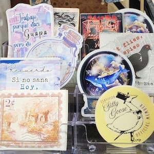 May include: A clear plastic organizer filled with various stickers. The stickers feature illustrations of a whale, a goose, and other designs. Some stickers have text in Spanish and English.