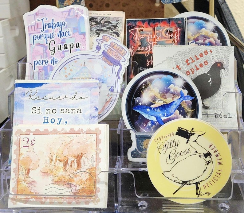 May include: A clear plastic organizer filled with various stickers. The stickers feature illustrations of a whale, a goose, and other designs. Some stickers have text in Spanish and English.
