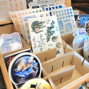 May include: Wooden organizer box filled with stickers and stationery. Stickers feature botanical illustrations, watercolor designs, and text. The box is divided into compartments, with a metal latch and handle.