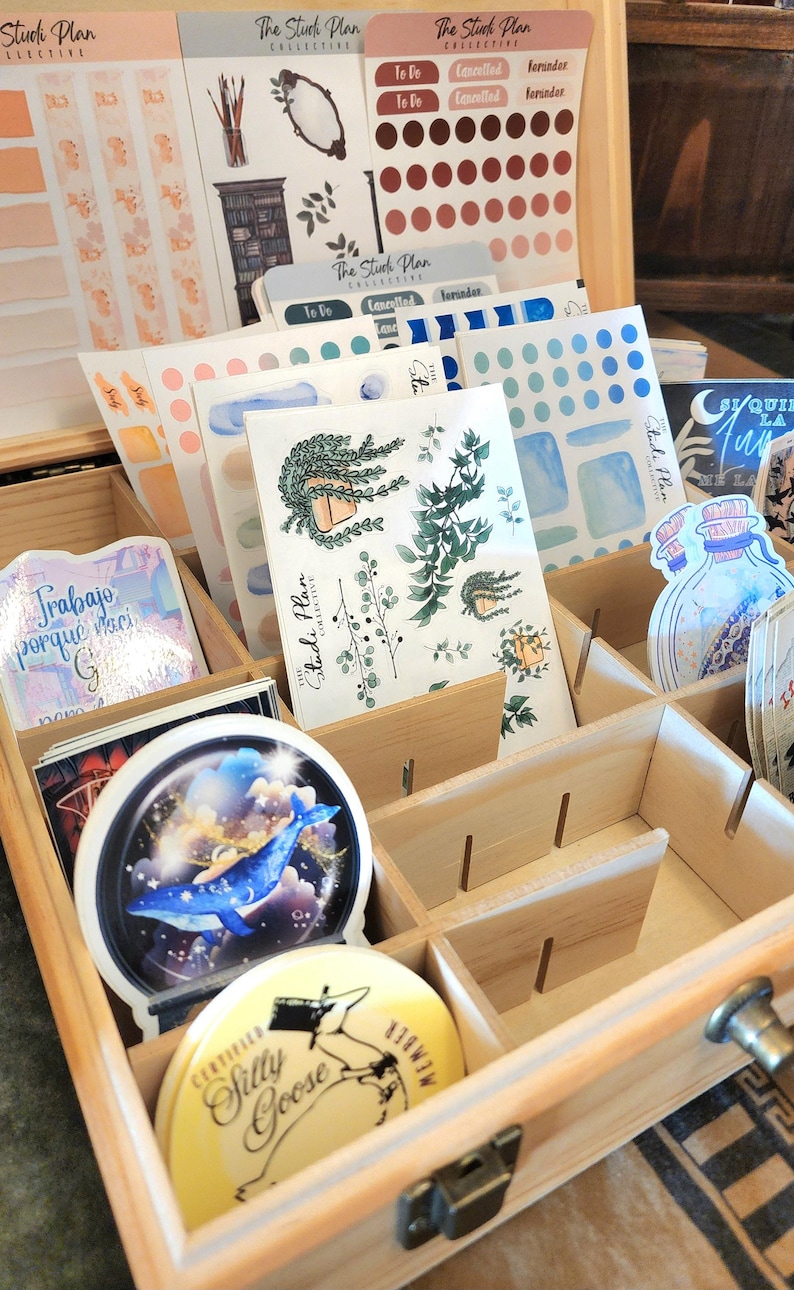 May include: Wooden organizer box filled with stickers and stationery. Stickers feature botanical illustrations, watercolor designs, and text. The box is divided into compartments, with a metal latch and handle.