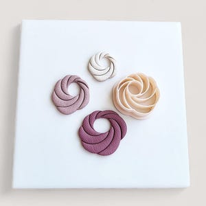 May include: Four abstract, spiral-shaped objects in shades of pink, beige, and purple are arranged on a white surface. The objects have a textured appearance, with ridges and curves. The arrangement is symmetrical, with the objects evenly spaced.