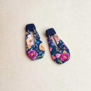 May include: A pair of floral earrings. The earrings feature a dark blue rectangular top and a teardrop-shaped body. The body of the earrings is decorated with a floral pattern in shades of pink, yellow, and white on a dark blue background.