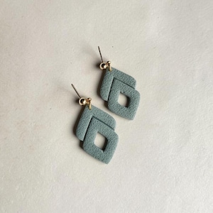 May include: A pair of light blue, textured clay earrings. Each earring features a layered, geometric design with a diamond-shaped cutout. The earrings have gold-colored ear wires and are displayed on a neutral background.