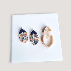 May include: Two floral pattern earrings with a dark blue top section. Two light pink earring-shaped cookie cutters are also present. The earrings have a gold-coloured hook. The floral pattern includes orange, blue, and white flowers.