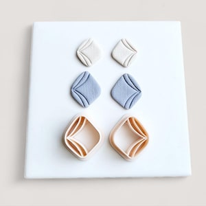 May include: Three pairs of diamond-shaped polymer clay earrings in cream, light blue, and peach. The earrings are displayed on a white surface, highlighting their geometric design and textured surfaces.