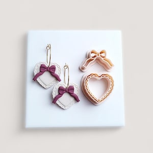 May include: An assortment of heart-shaped earrings and cookie cutters. The earrings have white heart pendants with scalloped edges, purple bows, and gold hoops. The cookie cutters are shaped like a bow and a heart.