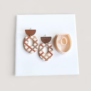 May include: Geometric earrings with a brown wooden top and a cream and brown patterned oval shape. A matching clay cutter is next to the earrings. The earrings and cutter are on a white surface.