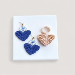 May include: A pair of blue and white patterned earrings with gold-colored posts and a matching clay cutter. The earrings have a layered, wavy design in a deep blue color, with a white and blue floral pattern at the top.