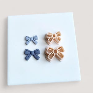 May include: Four bow-shaped objects in two different colours are arranged on a white surface. Two bows are a light blue, and two are a light tan colour. The bows are small and appear to be made of a solid material.