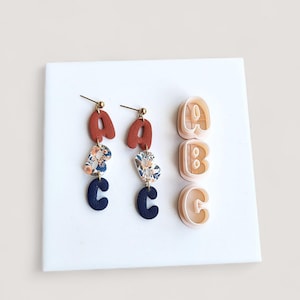 May include: Handmade earrings with the letters A and C, featuring a floral pattern and dark blue accents. The earrings are displayed with letter molds of A, B, and C.