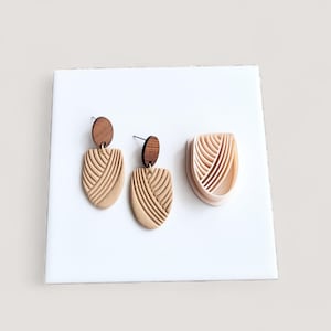 Peut inclure: Une paire de boucles d'oreilles beiges avec un dessus en bois et un emporte-pièce assorti. Les boucles d'oreilles ont un motif géométrique texturé. L'emporte-pièce a la forme des boucles d'oreilles. Les boucles d'oreilles sont sur une surface blanche.