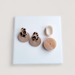 May include: Two pairs of beige and leopard print earrings with matching clay cutters. The earrings have a circular design with concentric rings. The leopard print is brown and black. The cutters are oval and circular.