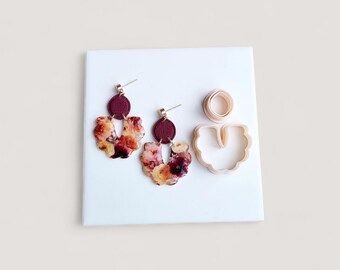May include: A pair of floral-patterned earrings with gold-colored posts and burgundy accents. The earrings are paired with a matching clay cutter set, including a circle and a U-shaped cutter, all on a white surface.