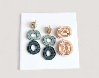 May include: Earrings with a gold leaf design and three stacked, open-circle shapes in shades of green. Two additional circular shapes in a light peach color are also present. The earrings are displayed on a white surface.