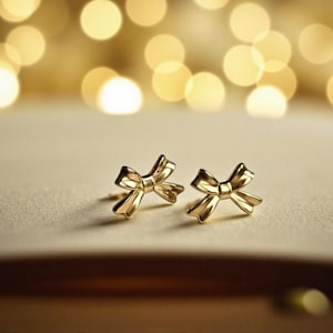 May include: A pair of gold-coloured bow-shaped stud earrings. The earrings are set on a light-coloured surface, with a blurred background of golden bokeh lights. The earrings are small and delicate, with a polished finish.