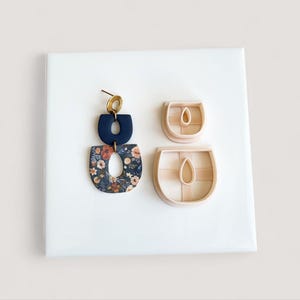 May include: A pair of handmade earrings with a gold-colored ear wire and two U-shaped components. The top component is navy blue, and the bottom is a floral pattern on a navy background. Two U-shaped clay cutters are also shown.