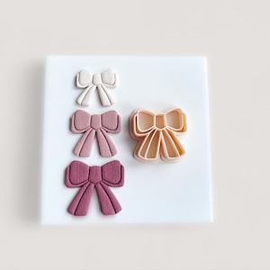 May include: An assortment of bow-shaped cookie cutters and finished bows in various shades of pink and cream. The cutters are made of a light peach-colored plastic. The finished bows are arranged on a white surface, showcasing the different colors and textures.