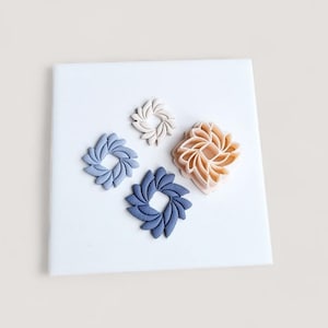 May include: Four decorative, floral-shaped objects in varying sizes and colors. The colors include light blue, dark blue, beige, and peach. The objects are arranged on a white square surface, creating a visually appealing arrangement.