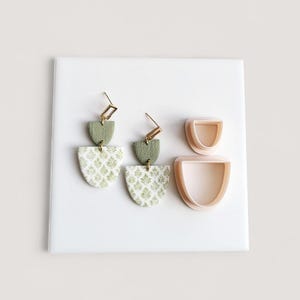 May include: A pair of handmade earrings with a gold-colored hook and a two-tiered design. The top tier is olive green, and the bottom tier is white with a green floral pattern. Two matching pink, semi-circular clay cutters are also present.