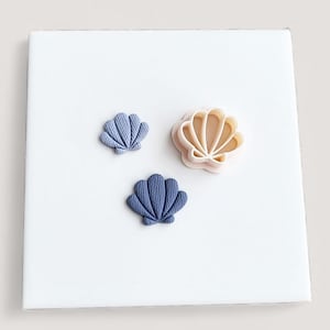 May include: Three seashell-shaped cookie cutters. Two are blue, and one is beige. The blue cutters have a textured surface, while the beige one has a smooth finish. The cutters are arranged on a white surface.