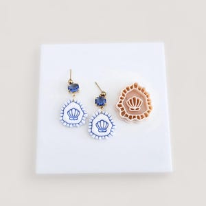 May include: A pair of blue and white seashell earrings with gold-colored posts and a matching seashell-shaped stamp. The earrings feature a blue gemstone at the top and a seashell design on the bottom. The stamp is a light brown color.