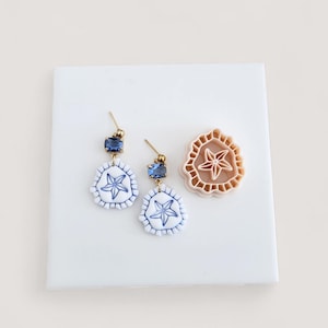 May include: A pair of earrings with a blue and white starfish design, gold-coloured posts, and blue rectangular gems. A matching starfish-shaped clay cutter is also present, showcasing a detailed starfish pattern. The earrings and cutter are on a white surface.