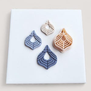 May include: Four polymer clay cutters in a teardrop shape with a detailed, layered design. Two are a muted blue, one is beige, and the other is a light peach color. The cutters are arranged on a white surface.