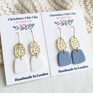May include: Two pairs of handmade earrings. One pair has white clay shapes, the other blue. Both have gold-coloured, openwork oval accents. The cards read "Christina's Chic Clay Handmade, Handmade In London."